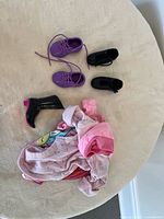 Photo displaying doll shoes and pink doll clothing items laid out on a cream surface.
