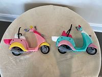 Top-down view of two Barbie Vespa scooters placed side by side showing colors and accessory details.