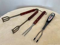 Photo showing two metal BBQ tongs and a BBQ fork with wooden handles, alongside a digital meat thermometer with dual metal probes and control buttons