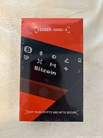 Front view of sealed Ledger Nano X box with red and black design and Bitcoin icons.