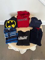 Photo showing six boys sweaters and sweatshirts arranged on a table, featuring brands Nike, Roots, and Batman in sizes 6-8
