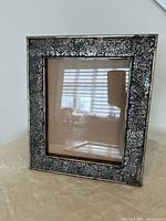 Rectangular brown and black textured glass picture frame with reflective glass cover, empty, showing cardboard back placeholder. Background shows reflection of window blinds and room.
