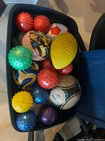 Photo showing a large black plastic bin filled with a variety of small balls including sensory spiked balls, a yellow small football, and several soccer balls in orange-white, blue-white, and black-white color patterns. Some balls show signs of use.