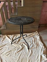 Black round glass outdoor side table with three metal legs on a cloth, placed indoors by wooden stairs.