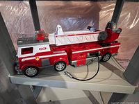 Front and side view of the large red and white toy fire truck placed on a plastic shelf, showing details of the cabin, ladder, wheels, and attached hose.