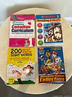 Four educational books laid out on a beige circular fabric surface, viewed from above. Books include a Grade 1 Canadian Curriculum book, Scholastic Reading & Math workbook, 200 Must Know Sight Words workbook, and a Paw Patrol sticker book.