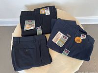 Three pairs of folded men's pants, two navy blue and one black, displayed on a round table with tags visible.