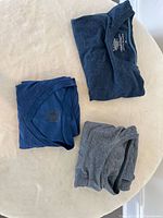 Top-down view of three folded men's t-shirts. Two are blue, and one is grey. The shirts are laid out on a light surface for clear visibility.