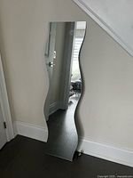 Full-length mirror with wavy sides standing against a wall near a doorway, reflecting part of the adjacent room.