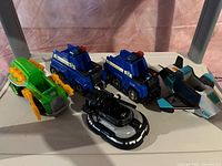 Photo showing three Paw Patrol character toy vehicles: two Chase police cars and one Everest snow vehicle, on a flat surface with a brown background, detailing colors and features of the vehicles.