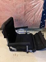 Side angled view of black portable seat showing padded backrest, seat cushion, armrest, and partially extended footrest on concrete floor.