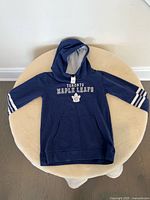 Blue kids hoodie laid flat on a round beige stool against a white wall. Front features Toronto Maple Leafs text and logo, sleeves have white stripe details. The interior of the hood is gray.