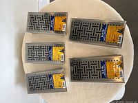 Top-down photo of five unopened metal vent grates arranged on a round beige surface, showing their full front face and packaging labels.