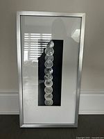 Framed modern wall artwork with a row of ten circular textured elements on a black background inside a silver metallic frame.