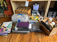 Wide view showing art supplies arranged on floor: plastic art supply boxes, sketchbooks, colored pencils, wooden easel, and other containers.