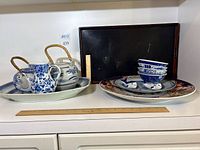 Two teapots on white oval tray next to large round floral platter, rice bowls, chopstick rests and small pitcher