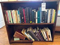 Photo showing two wooden shelves filled with assorted books in varying colors and conditions, with some books stacked horizontally and others vertically.