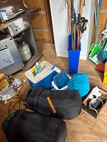 Photo showing black sleeping bags, blue and white coolers, assorted walking sticks in blue container, and other camping items scattered around a wooden floor.