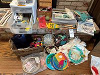 Overview of craft supplies on brick hearth and surrounding area, showing plastic drawers, baskets with yarn, decorative embroidery plates, tins with colored pencils, and paint tubes