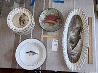 Top view of 12 ocean-themed appetizer plates with fish images, Italian porcelain bowl with fish, Williams Sonoma bowls, and oval serving platter with seal design and floral edge.