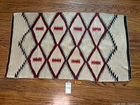 Full view of the rectangular hand woven dual sided rug displaying red diamond geometric patterns with black and grey accents on a beige background.