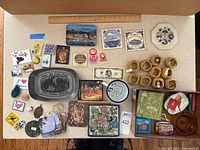 Photo showing entire assortment of trivets, coasters, magnets, napkin rings, pins, keychains, and decorative plates spread on a table with ruler for scale.