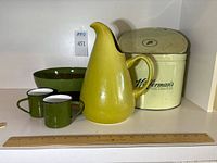 View of green-yellow ceramic pitcher, pair of green enameled mugs, enameled bowl, and vintage metal storage canister together on shelf with ruler for size reference