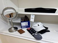 Wide view of lot items including face mirror with ring light, Birkenstock box, wallet, stack of black velvet hangers, black gloves, socks, and Mickey Mouse cap on white shelf.