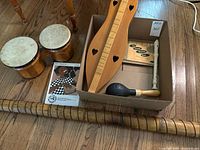 Overview of pair of wooden bongos, cardboard box containing dulcimer, recorder, shaker, music book, and wooden tube instrument