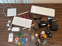 Overview of lot showing two Apple wireless keyboards, Apple Magic Mouse, HP 35s calculator in case, assorted pins, keychains, and accessories arranged on a table.