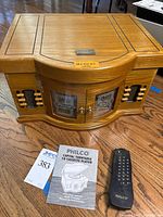 Photo of the closed wooden cabinet housing the media player with two front glass doors featuring Philco logos, remote control, and manual placed on the floor.