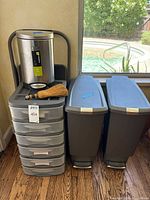 Full view of the lot showing two tall plastic waste bins with blue lids, small metal waste bin, whisk broom, and six drawer plastic storage unit