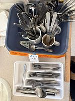 Wide view of mixed flatware in a blue tub including knives, forks, spoons and other utensils organized in mugs, and plastic containers beside them.