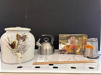 Wide view showing all four main items: ceramic water dispenser with butterfly design, steel kettle, boxed glass tea infuser, and coffee grinder.