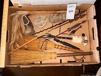 Top-down view of all items in a cardboard box showing the crafts including brooms, carved faces, spears, shoe, and other wood carvings.