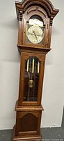 Full view of grandfather clock showing wooden case, glass door with weights and pendulum, clock face with black numerals and hour/minute hands, decorative arched top.