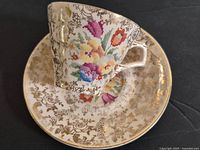 Close-up angled photo of the teacup on the saucer showing floral cross-stitch sampler pattern and gold filigree