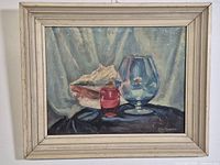 Framed oil painting showing the entire piece including the white rustic wooden frame and the painted still life of a conch shell, red glass, and large blue glass.