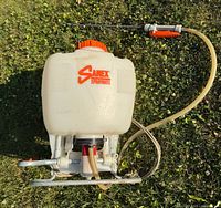 Top view of white Sanex Spraymate backpack sprayer with orange cap and attached spray wand and hose showing pump lever and port fittings.
