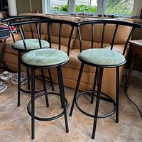 Three black metal frame bar stools with fabric covered round seats. They have a curved backrest with vertical supports, standing on four legs with circular footrests.