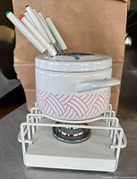 White ceramic fondue pot on white metal burner stand, with 10 forks inserted into the pot lid. Pot has pink geometric pattern and side handle.