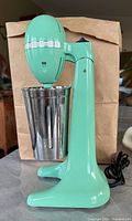 Front-left angle view of the green Hamilton Beach milkshake maker with stainless steel cup, showing power cord and stable pedestal base.