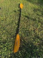 Full view of the 84-inch black shaft paddle with bright orange offset blades on grass.