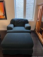 Photo of blue armchair with cushioned armrests and matching ottoman placed in a room with wooden flooring and light from a window behind.