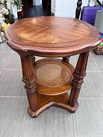 Round wooden end table with decorative top and woven cane lower shelf, carved legs and scalloped edges.