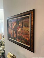 Framed landscape painting showing a countryside village scene with buildings, trees, and fields in warm colors, displayed hanging on a white wall.