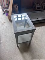Glass coffee table standing on a concrete floor, shows rectangular glass inset surrounded by white painted wooden frame with minor wear.