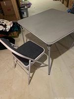 Grey folding table and folding chair set shown from an angle, displaying table surface and chair padded seat.