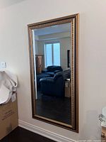 Full view of the large rectangular mirror standing against a wall, reflecting a room with black leather furniture and windows. The frame is bronze-colored with textured design.