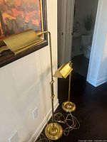 Pair of two adjustable brass floor lamps with round weighted bases and cylindrical shades, shown standing next to a wall.